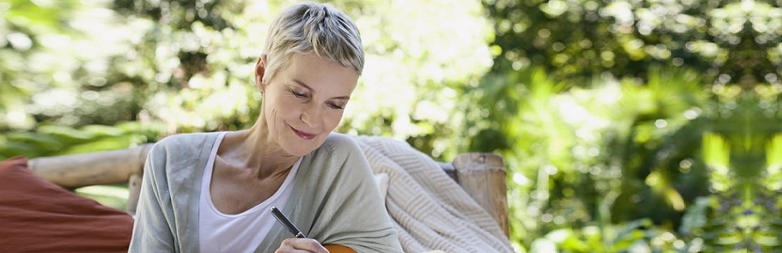 A mature woman is seated on a bench in an outdoor space. She is smiling and writing in a notebook with a pen.